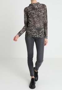 Leopardmönstrad långärmad topp av genomskinligt material, matchad med grå slim-fit byxor och svarta snörskor. Enkel bakgrund.
