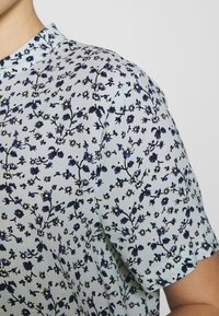 Bluse mit floralen Mustern in Himmelblau mit marineblauen und grünen Details. Verfügt über einen Kragen und kurze Ärmel, aus leichtem Stoff gefertigt.