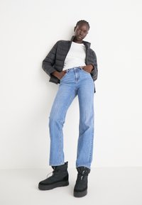 Chaqueta acolchada negra, camisa blanca, jeans de tiro alto azul y botas negras. Texturas notables en la chaqueta y denim en contraste.