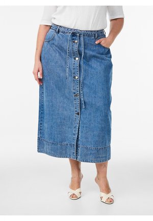 Nainen, jolla on midimittainen sininen farkkuhame, jossa on napit ja vyötäisillä solmittava vyö, valkoinen toppi ja valkoiset avovarvassandaalit korolla.