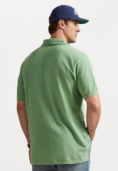 Άνδρας με πράσινο κοντομάνικο πόλο μπλουζάκι και μπλε καπέλο του μπέιζμπολ, κοιτάζοντας στο πλάι με τα χέρια στις τσέπες, μπροστά σε λευκό φόντο.