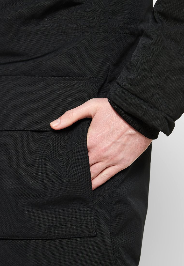 Abrigo negro con tejido texturizado y un bolsillo lateral. Una mano está colocada en el bolsillo, mostrando el diseño de la prenda.