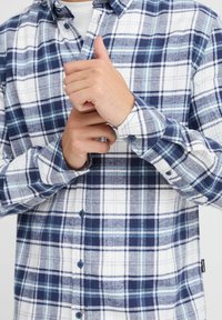 Camicia a quadri blu e bianca in tessuto flanella con design a bottoni. Presenta strisce alternate spesse e sottili, polsini arrotondati e bottoni.