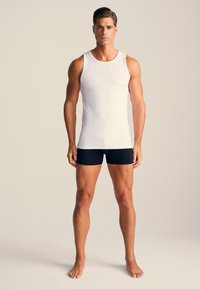 White sleeveless tank top and black boxer shorts modeled by a male figure. The fabric appears smooth with a fitted design.