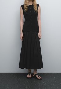 Zwarte mouwloze top met knopen, gecombineerd met een lange zwarte geplooide rok en platte zwarte sandalen. Glad materiaal, minimalistisch ontwerp.