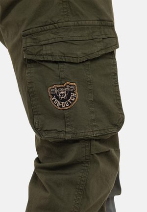 Gros plan sur la jambe d'un pantalon cargo vert olive avec une poche à rabat et un écusson logo Von Dutch marron et noir.