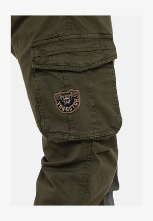 Gros plan sur la jambe d'un pantalon cargo vert olive avec une poche à rabat et un écusson logo Von Dutch marron et noir.