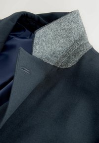 Gros plan du col d'une veste bleu foncé avec une doublure intérieure en feutre gris et des coutures visibles sur un fond clair.