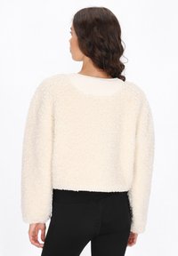 Maglione cropped di colore crema, morbido e fluffy, con finitura texturizzata, spalle a caduta e scollatura rotonda sul retro. Indossato sopra leggings neri.