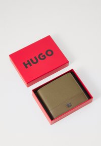 Green textured leather wallet in a red HUGO box, featuring a metal logo and clean design. Box has a red exterior with black lettering.