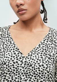 Μαύρη και λευκή μπλούζα με πουά και V-λαιμόκοψη, φτιαγμένη από απαλό ύφασμα, με κλείσιμο με κουμπιά μπροστά. Περιλαμβάνει διαφορετικά μεγέθη πουα και λεία υφή.