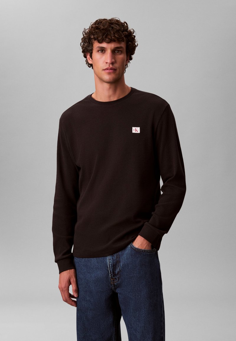 Czarna koszula z długim rękawem o prążkowanej fakturze, z okrągłym dekoltem i małą białą naszywką z logo. W zestawieniu z niebieskimi dżinsami, ręce w kieszeniach.