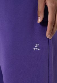 Lilla joggebukser laget av myk stoff, med en liten hvit brodert logo på siden. Glatt tekstur med en tettsittende design.
