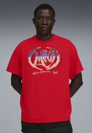 Młody mężczyzna stojący z rękami za plecami, ubrany w czerwony T-shirt z graficznym nadrukiem i stylizowanym tekstem oraz czarne spodnie o fakturze.