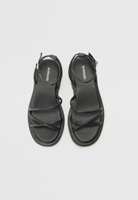 Sandales noires avec des lanières croisées, talon ouvert et boucle à la cheville. Texture lisse, bout arrondi, semelle plate, marquage minimal sur la semelle intérieure.