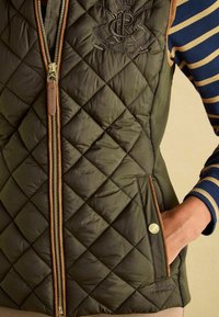 Prešívaná olivovozelená vesta s predným zipsom, hnedými koženými detailmi a vyšitým logom. Obsahuje bočné vrecká a má hladkú textúru.