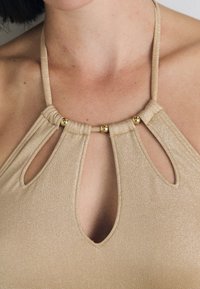 Beige halter nyakú top, amely kivágott részletekkel és arany tónusú díszítéssel rendelkezik. Az anyaga enyhe fényességgel és sima textúrával bír.