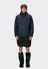 Marineblå vandtæt jakke med hætte, udstyret med lynlås foran, elastisk talje og i kombination med sorte shorts og blanke sorte sko.