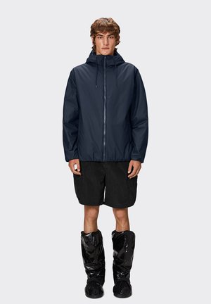 Giacca waterproof navy con cappuccio, dotata di zip frontale, vita elasticizzata, abbinata a pantaloni corti neri e calzature nere lucide.