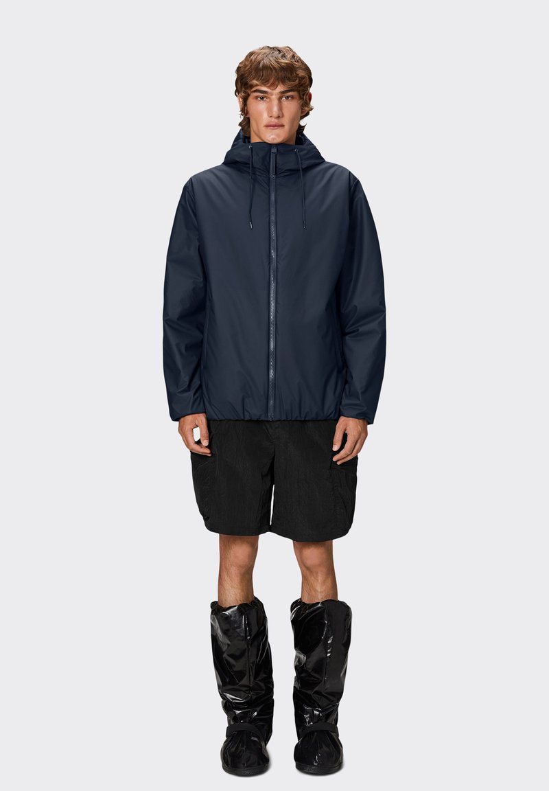 Marineblå vandtæt jakke med hætte, udstyret med lynlås foran, elastisk talje og i kombination med sorte shorts og blanke sorte sko.