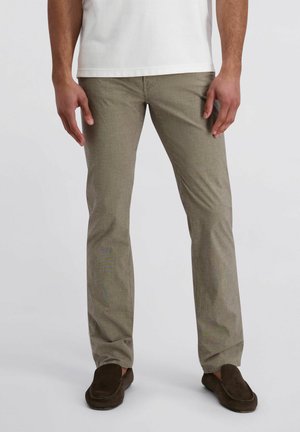 Mann trägt beige gerade geschnittene Hose, braune Wildleder-Loafer und weißes Hemd, steht vor hellem, einfarbigem Hintergrund.