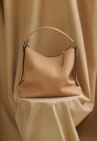 Pfirsichfarbene Hobo-Tasche aus Leder mit abgerundeter Form, einem Reißverschluss oben und einem einzelnen Griff. Strukturiertes Oberflächenmaterial und dezente tonale Akzente.