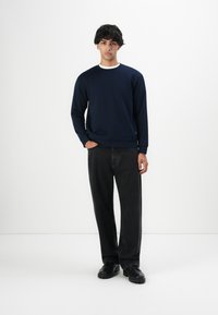 Joven con cabello rizado oscuro, vistiendo un sudadera azul marino, jeans negros y zapatos negros, de pie con una mano en el bolsillo.