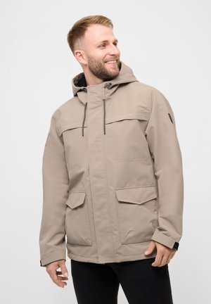 Hombre sonriendo y mirando de lado, vistiendo una chaqueta beige con capucha y bolsillos con solapa, y pantalones negros, sobre un fondo liso.