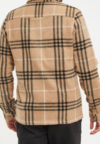 Personne portant une chemise en laine beige à carreaux avec un motif à damiers noir et blanc, associée à un pantalon noir, vue de dos.