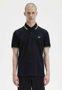 Polo shirt nera con colletto e polsini a righe dorate, con un piccolo logo ricamato sul petto. Materiale in cotone, maniche corte.