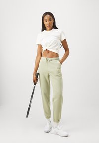 Light green sweatpants with elastic cuffs and drawstring waist, paired with a white knotted T-shirt. Holding a black tennis racket.