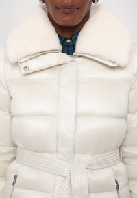 Pūdera jaka spīdīgā baltā materiālā ar mākslīgās ādas apkakli, jostas dizainu un kviltota tekstūra. Iespējas ietver sudraba aparatūru un sānu rāvējslēdzējums kabatas.