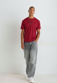 Κόκκινο βαμβακερό t-shirt με στρογγυλή λαιμόκοψη και σχέδιο λογότυπου, σε συνδυασμό με ανοιχτό γκρι άνετους τζιν και λευκά αθλητικά παπούτσια.