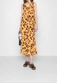 Top giallo-arancio senza maniche con stampa leopardata e volant, abbinato a una gonna coordinata. Clog marroni con borchie. Piccola borsa nera.