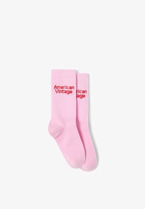Paire de chaussettes roses côtelées montantes avec le texte rouge "American Vintage" sur la partie supérieure du mollet, posées à plat sur un fond blanc.