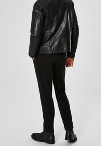 Man som bär en svart läderjacka, svarta byxor och svarta stövlar, står med händerna avslappnat ned längs sidorna, vänd bort från betraktaren.