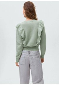 Svetlo zelena jopica z nabranimi šivi na ramenih, okroglim izrezom in prilegajočimi manšetami, v kombinaciji z svetlo sivimi hlačami visoke pasu.