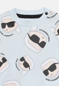 Material albastru deschis cu fețe de desene animate repetate, purtând ochelari de soare negri și păr alb, acompaniate de textul "KARL LAGERFELD."