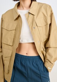 Khaki oversize kurtka zapinana na guziki z dwoma dużymi kieszeniami na piersiach, noszona na jasnym, krótkim topie i niebieskich sportowych szortach.