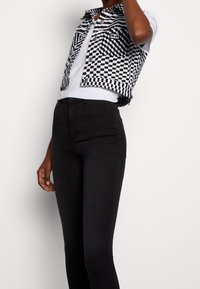 Persona vestida con pantalones ajustados negros de cintura alta, camiseta blanca y un chaleco recortado de cuadros en blanco y negro, de pie contra un fondo blanco.