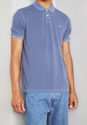 Mężczyzna ubrany w wyblakłą, niebieską koszulkę polo z krótkim rękawem i guzikami oraz jasnoniebieskie dżinsy, stojący na jasnym tle.