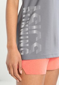 Maglietta sportiva grigia con stampa argentata traforata "RUNNING" sul lato, abbinata a pantaloncini color corallo con tessuto strutturato e orlo inclinato.