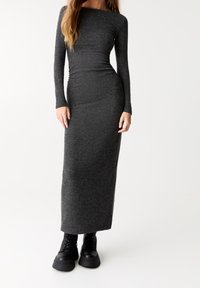 Robe grise moulante à manches longues, confectionnée dans un tissu lisse et extensible, dotée d'un col bateau et d'une coupe longue jusqu'au sol, associée à des bottes à plateforme noires.