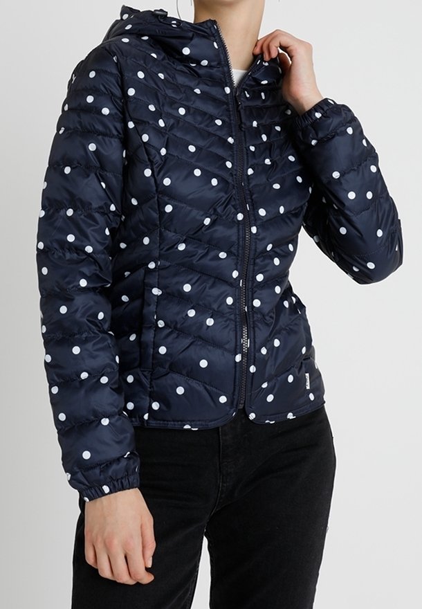 Veste matelassée bleu marine à pois blancs, fermeture éclair sur le devant, poignets élastiques et capuche attachée, portée avec un pantalon noir.