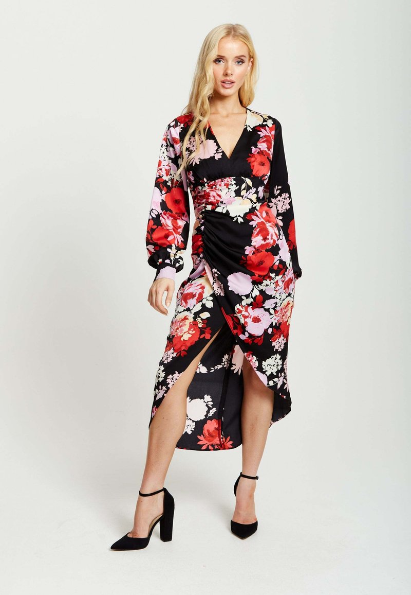 Black floral dress with long sleeves, featuring red and pink flowers, a wrap design, and side slit. Paired with black high-heeled shoes.