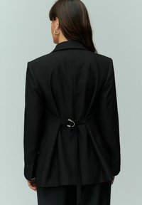 Sort blazer med strukturerede skuldre og skræddersyet pasform. Bemærkelsesværdige detaljer inkluderer en indsnævret ryg med en sølvspænde og klaplommer.