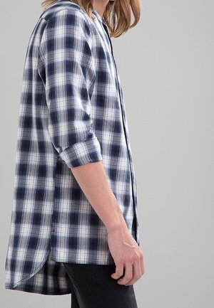 Personne portant une chemise à carreaux bleue et blanche à manches longues avec les manches retroussées, debout devant un fond gris uni.