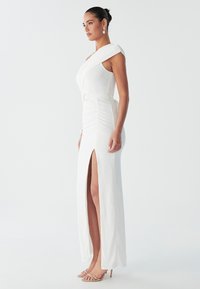 Weißes, einseitig schulterfreies Kleid mit Raffungen, seitlichem Schlitz und einem Gürtelakzent. Glatte Textur, knöchellang. Kombiniert mit hautfarbenen High Heels.