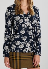 Námořnická květinová blůza s bílými květinovými vzory, V-štíhlý výstřih, dlouhé rukávy. Kombinováno s hnědou kostkovanou minisukní s žlutými akcenty.