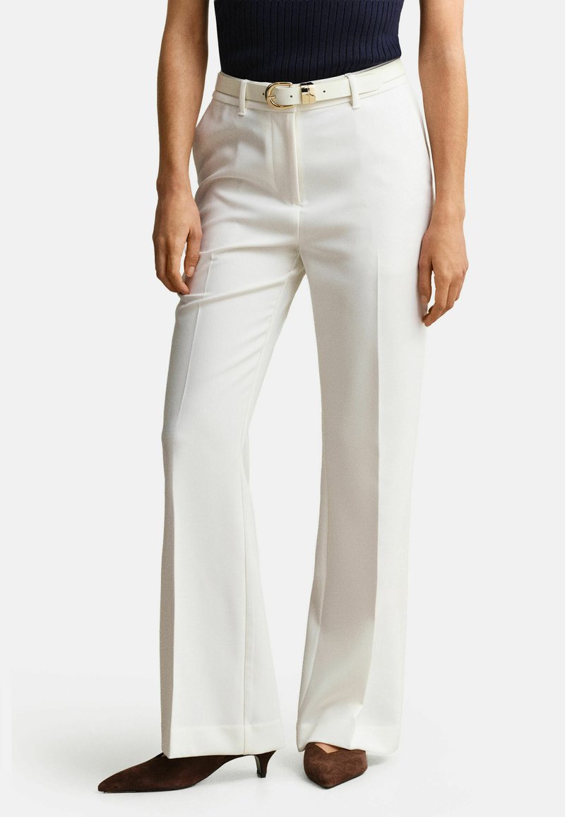 Vrouw draagt witte op maat gemaakte broek met een witte riem, zwart topje en bruine, puntige lage hakken, staand tegen een effen achtergrond.